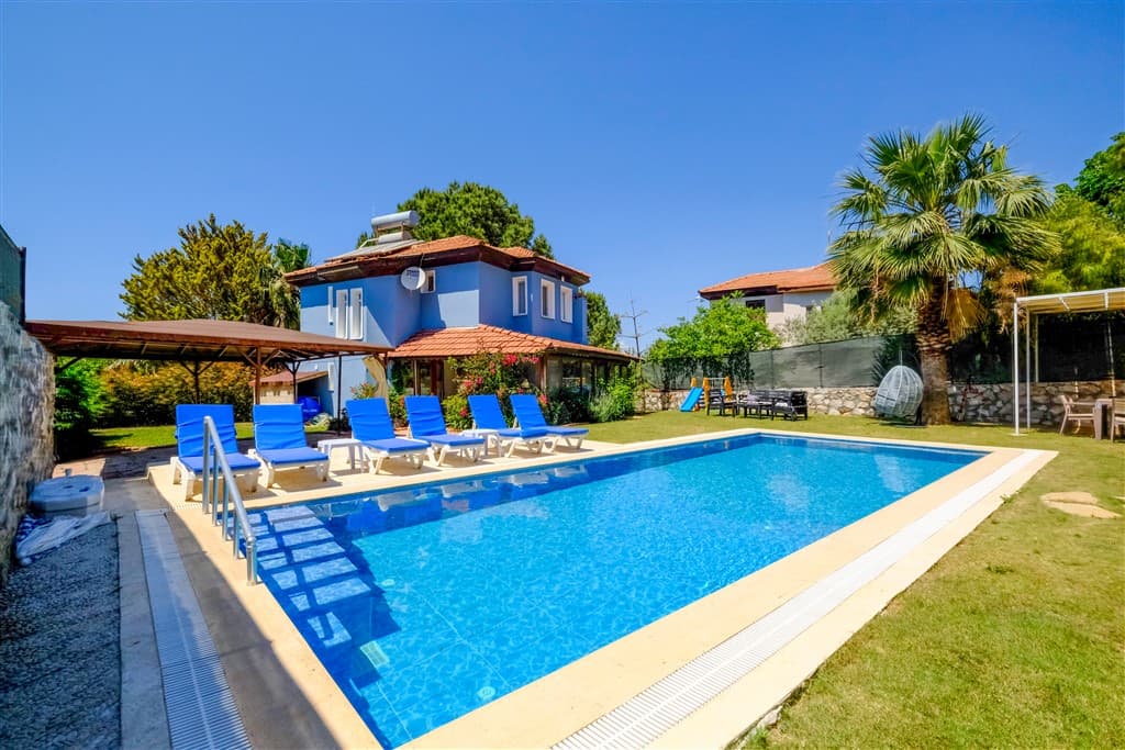 Bu emlak fotoğrafında, parlak güneş ışığı altında, masmavi bir havuza sahip geniş bahçeli mavi bir villa yer almaktadır. Havuz kenarında güneşlenme şezlongları, gölgelikli bir oturma alanı ve çocuk oyun grubu gibi keyifli yaşam alanları bulunmaktadır.